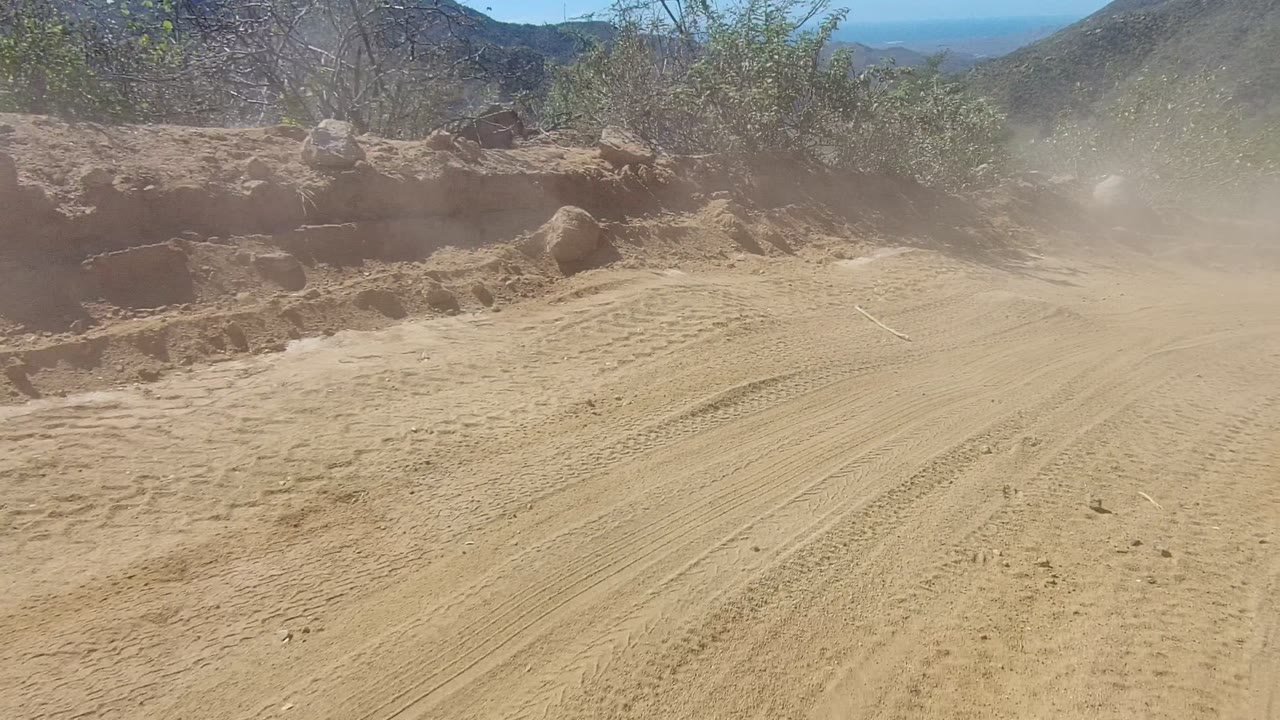 A dusty sandy road in Los Cabos, Baja California Sur, Mexico, shows tire tracks, scrub vegetation, rocky edges, distant hills, and no visible people.