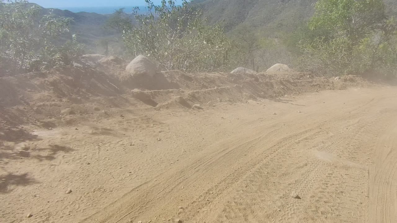 Dust cloud over a curving sandy road bordered by brush and rocks
