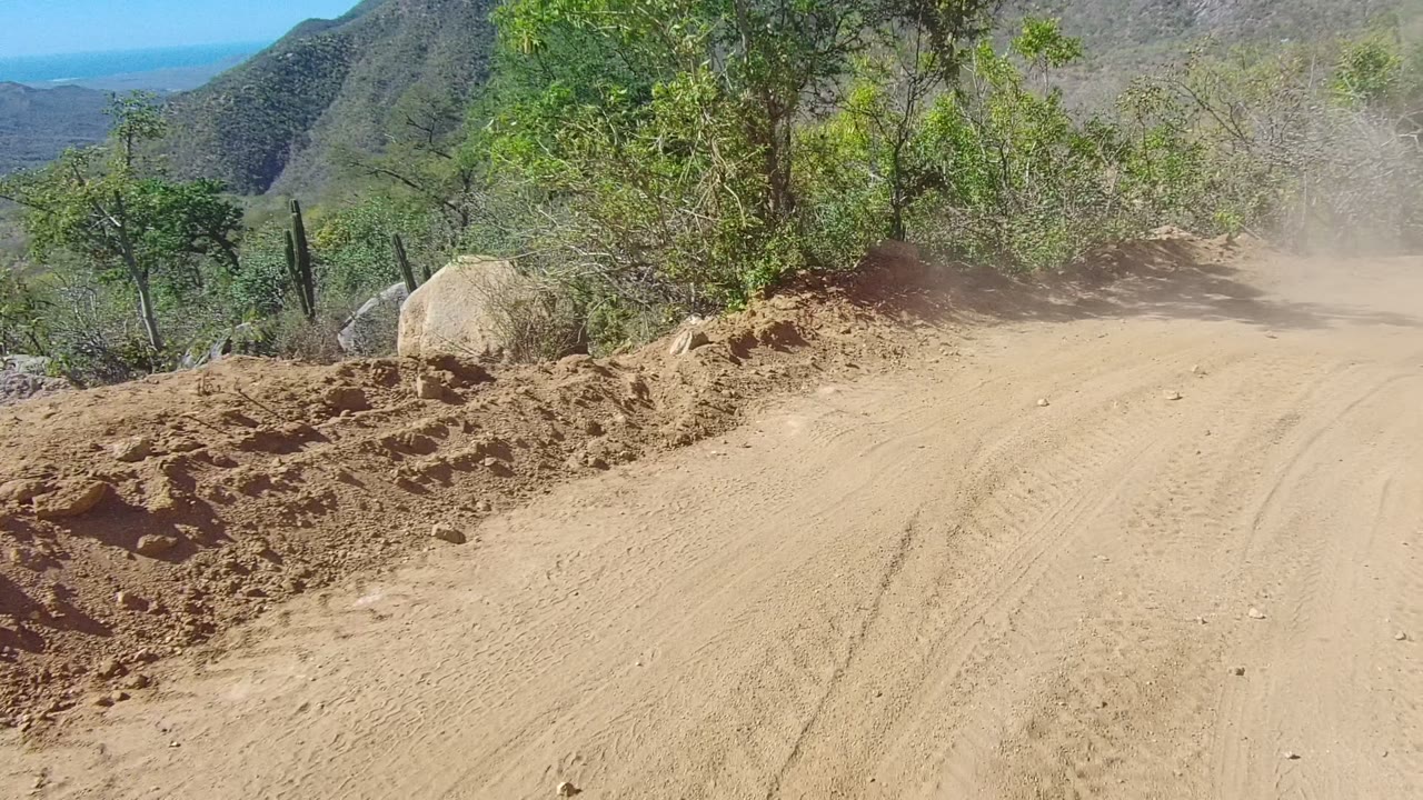 The Los Cabos dirt road curves past a rocky berm and brush, with mountain slopes and a blue distant horizon visible, and no visible people.