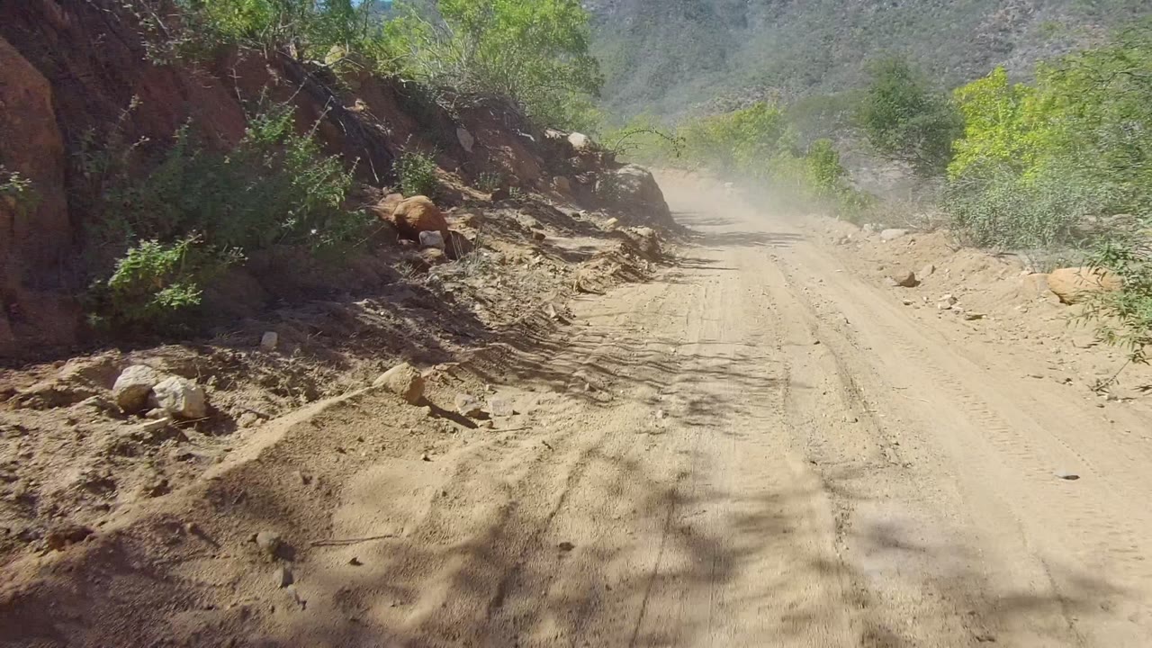 Dusty dirt road in Los Cabos, Baja California Sur curves past rocky edges and green brush, with no visible people in this opening frame.