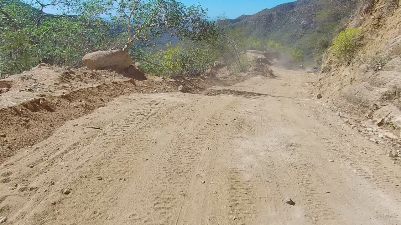 Opening frame of a dusty unpaved road in Los Cabos, Baja California Sur, with tire tracks, scrubby trees, rocky banks, distant hills, and no visible people.