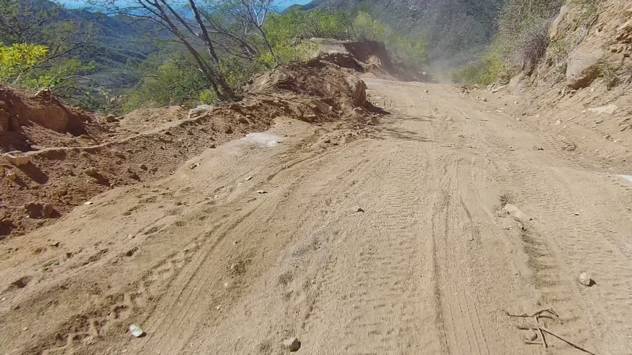 The road view opens slightly in Los Cabos, with dusty tire ruts, tree shadows across the track, brush on both sides, distant mountain ridges, and no visible people.