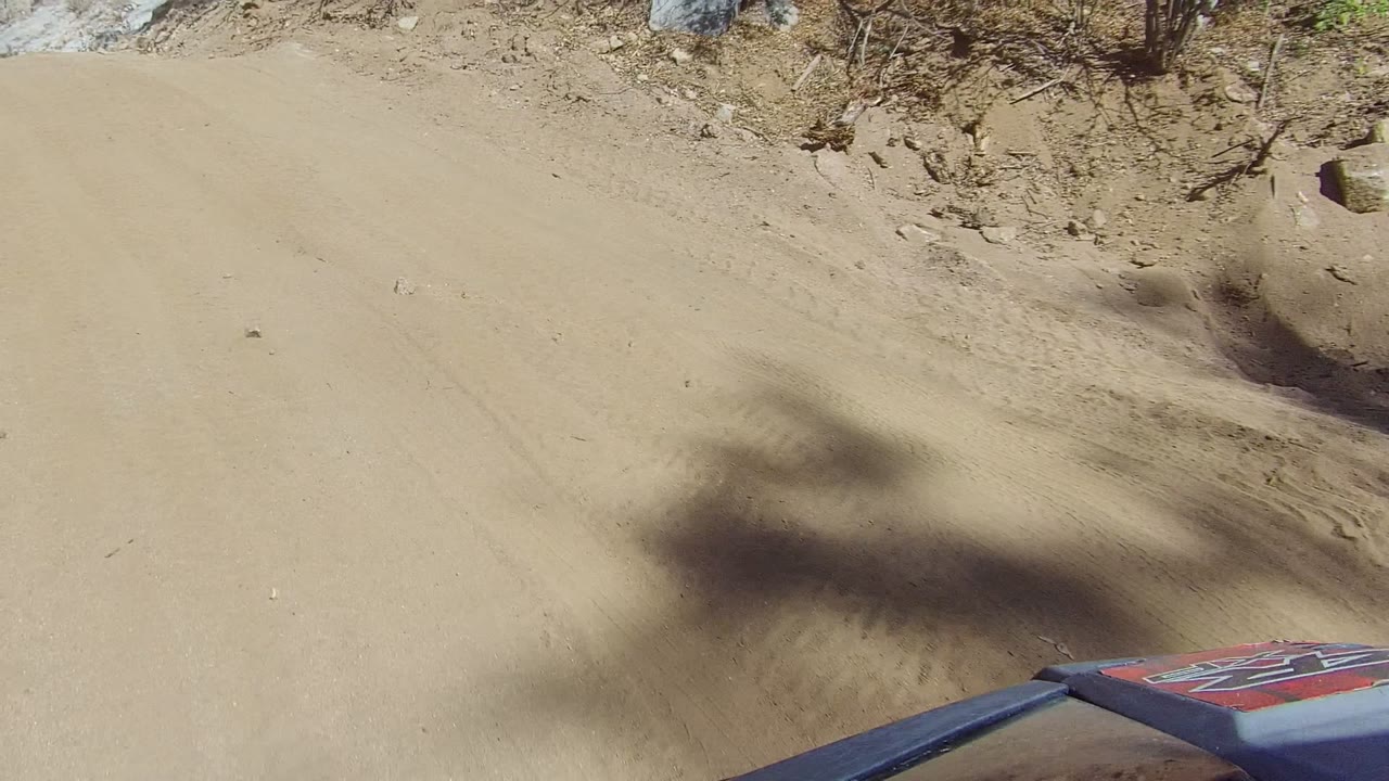 A partial vehicle hood or body panel appears at the lower right as the Los Cabos dirt trail curves past brush and rock, with no visible people.