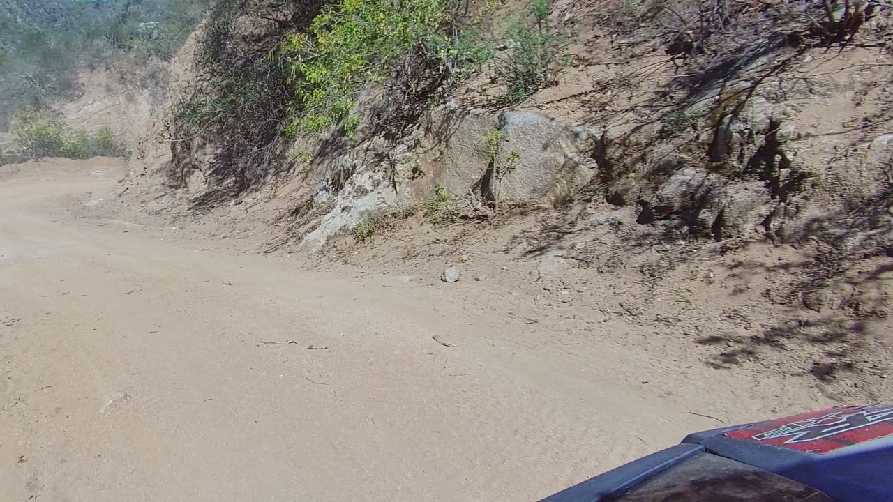 Forward view on a dusty Los Cabos dirt road beside a rocky bank and sparse brush, with a small vehicle hood edge visible and no visible people.