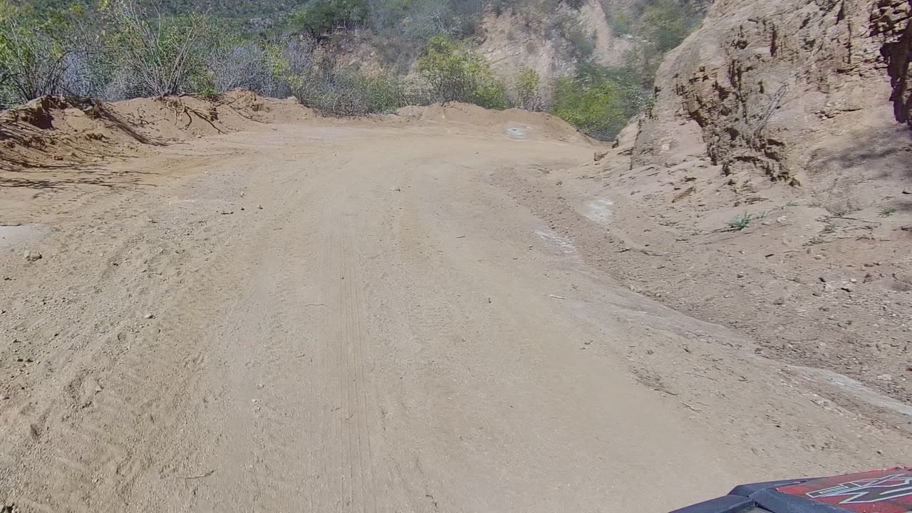 The Los Cabos trail continues toward a sandy rise, with brush along both sides, rocky cuts at the edges, a vehicle hood corner, and no visible people.