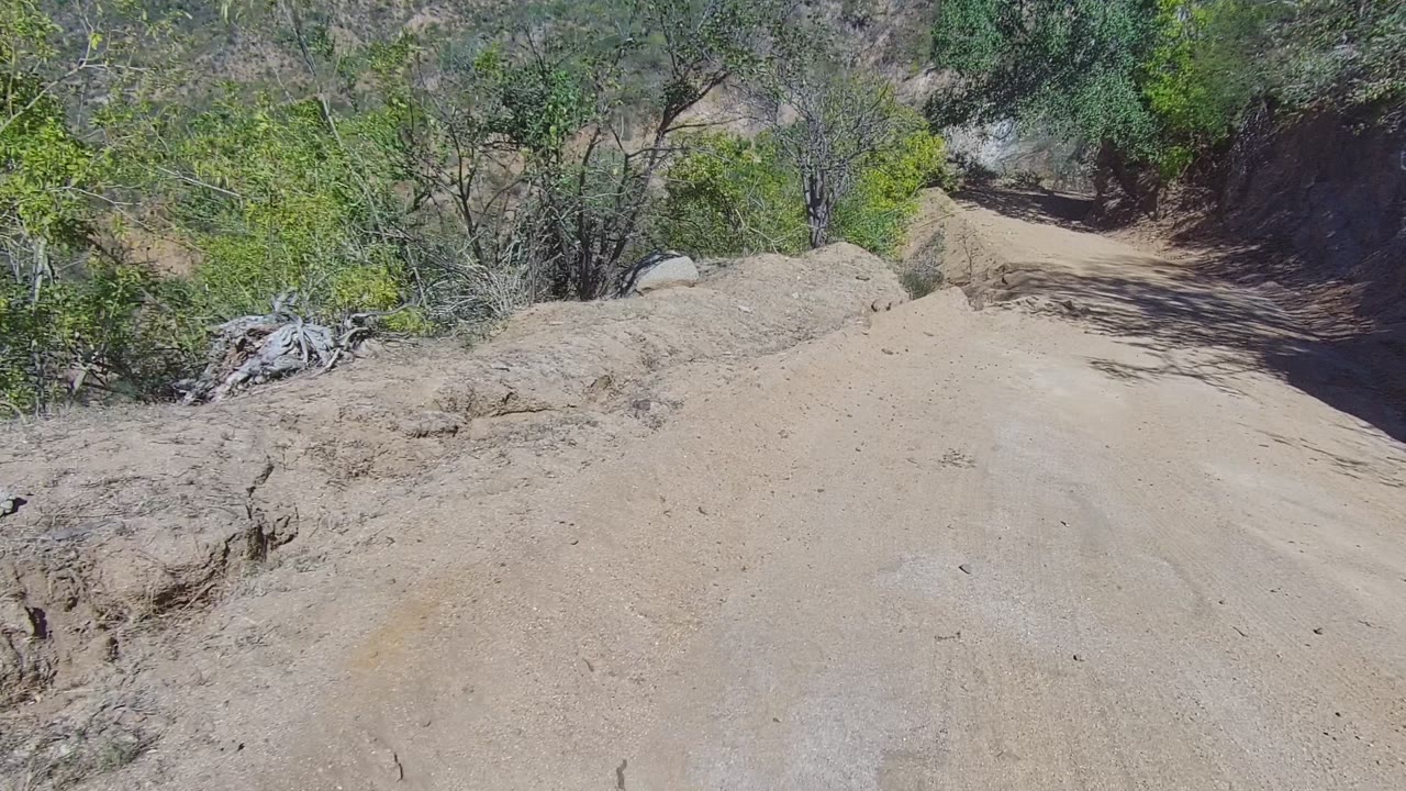 Forward view on a dusty dirt road in Los Cabos, Baja California Sur, with dry hillsides, scattered green trees, and no visible people at the start of the clip.
