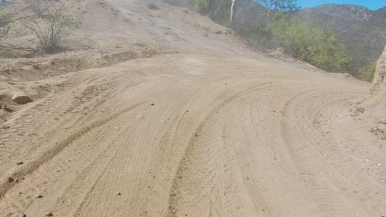 Low vehicle-view frame of a sandy dirt road with tire marks, dry banks, sparse shrubs, and blue sky in Los Cabos, Baja California Sur, with no visible people.