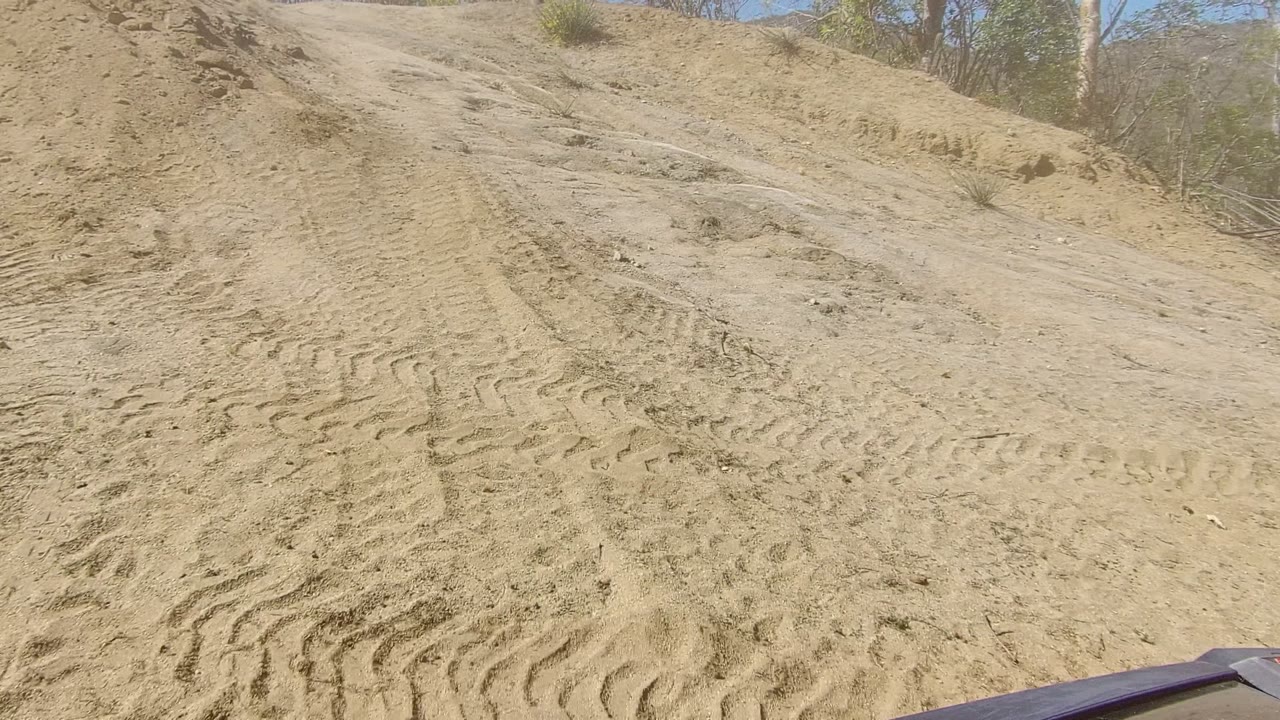 Sandy off-road slope with dense tire impressions, loose ruts, dry scrub, and the corner of a vehicle hood or body entering the lower right in Los Cabos, Baja California Sur, with no visible people.