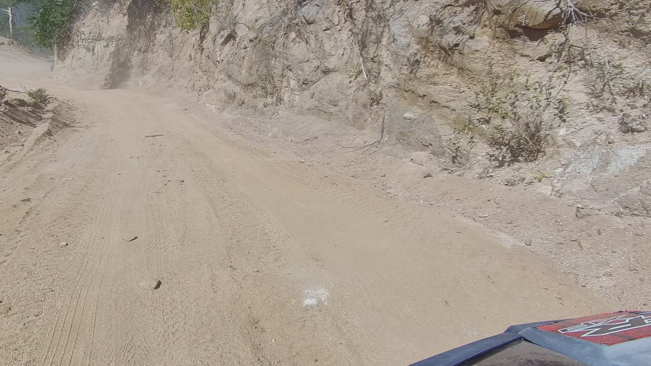 Forward view on a Los Cabos dirt road at the start of the clip, with tire tracks, eroded rock walls, sparse shrubs, a vehicle hood edge, and no visible people.