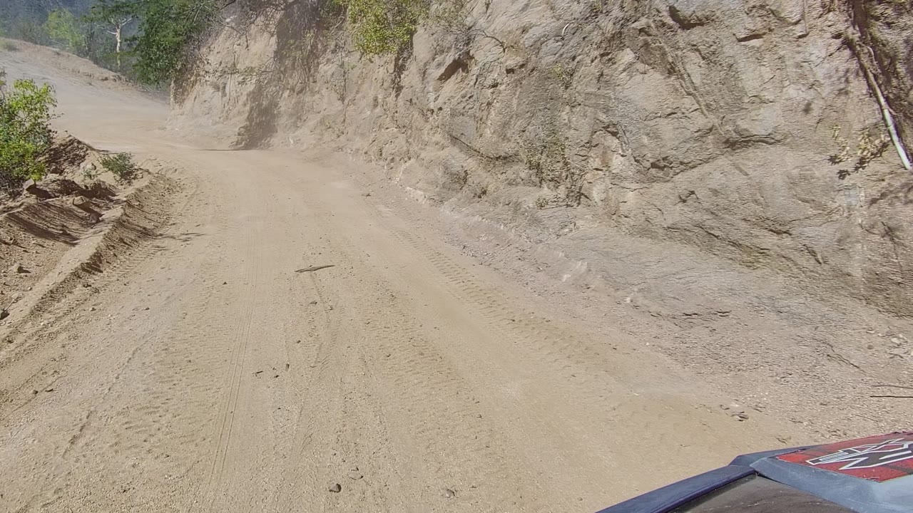 Narrow tan dirt road bordered by steep eroded rock and sparse greenery.