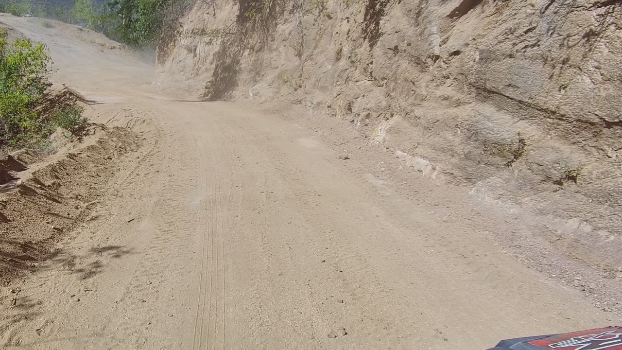 A bright midday view shows the Los Cabos dirt road bending past a close rocky wall, with dusty surface texture, faint tire tracks, scattered stones, and no visible people.