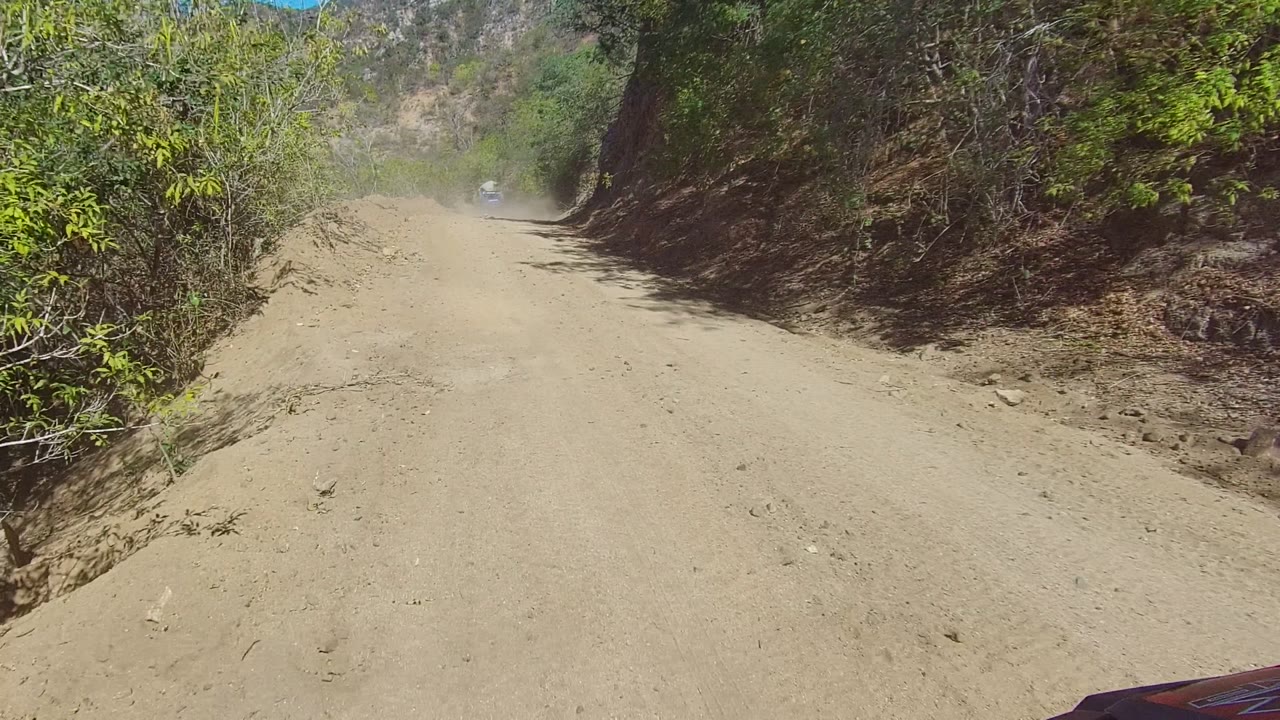 A forward view down a dusty dirt road in Los Cabos, Baja California Sur, with green brush on the left, a rocky bank on the right, distant hills, and no visible people.
