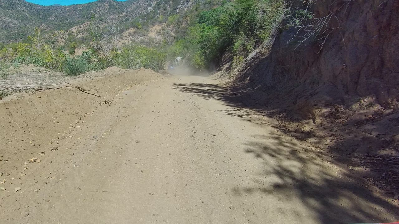A wider section of the Los Cabos dirt road shows a raised sandy berm at left, brush and reeds near the track, a rocky wall at right, distant mountains, dust ahead, and no visible people.