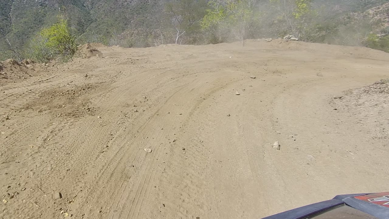 Wide dusty track in Los Cabos, Baja California Sur with a rock wall to the left, scattered green trees in the background, and no visible people.