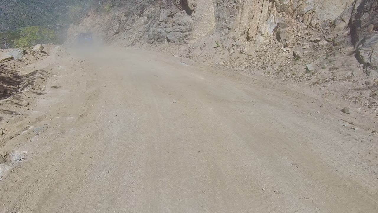A dusty dirt road in Los Cabos runs between tan rocky slopes, with a dust cloud ahead and no visible people at the start of the clip.