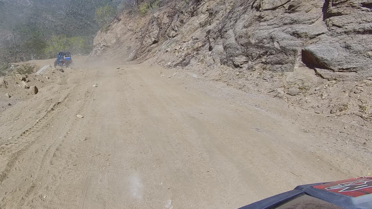 A blue vehicle appears ahead on the Los Cabos mountain dirt road, partly veiled by dust near a rocky bend, with no visible people.