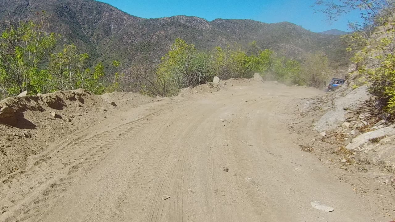 A dusty dirt road in Los Cabos, Baja California Sur curves through rocky dry terrain toward scrub-covered mountains, with a small blue vehicle barely visible ahead and no visible people.