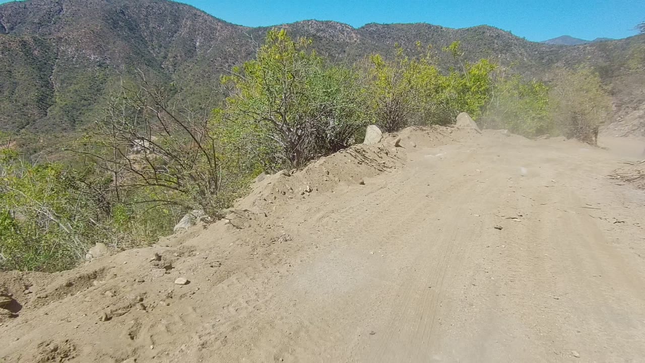 Wide dirt road bordered by brush and rocks with dust hanging over the route.