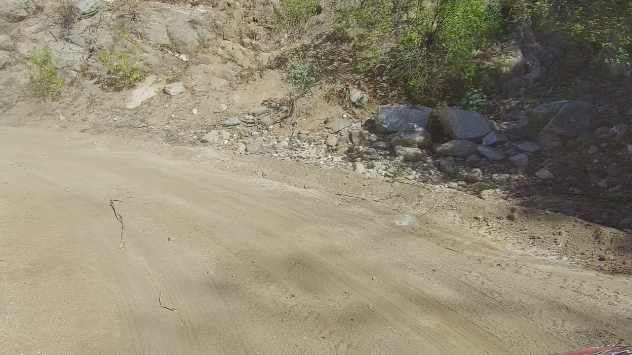 Empty dusty road in Los Cabos, Baja California Sur, Mexico, runs past a rock face and low shrubs under bright sun, with no visible people.