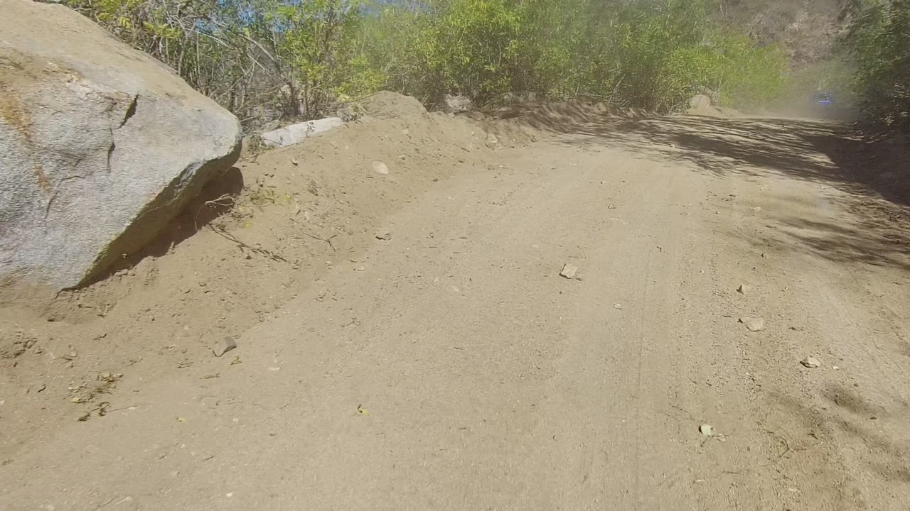 A sandy mountain path in Los Cabos passes a large boulder and scrubby green vegetation, with no visible people.