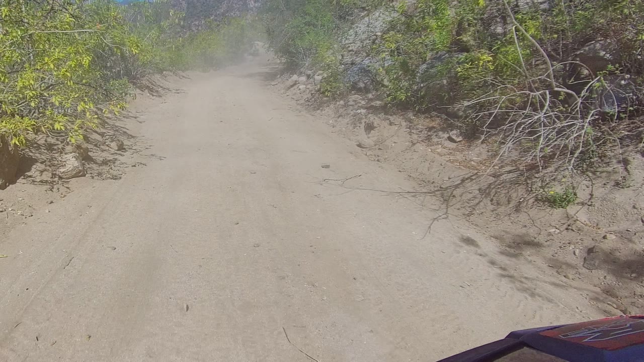 Sandy road beside brush and large rocks with dust ahead.
