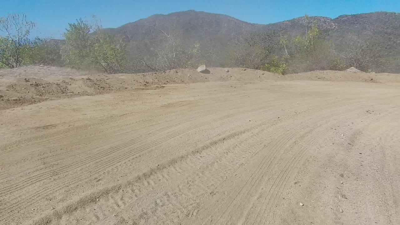 Wide view of a dusty dirt road in Los Cabos, Baja California Sur, with low scrub, distant mountains, bright blue sky, and no visible people.
