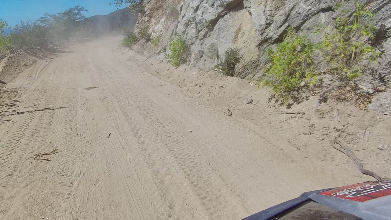 Start of a Los Cabos dirt-road drive, with sandy tire tracks, a rocky cliff edge, sparse shrubs, blue sky, and no visible people.