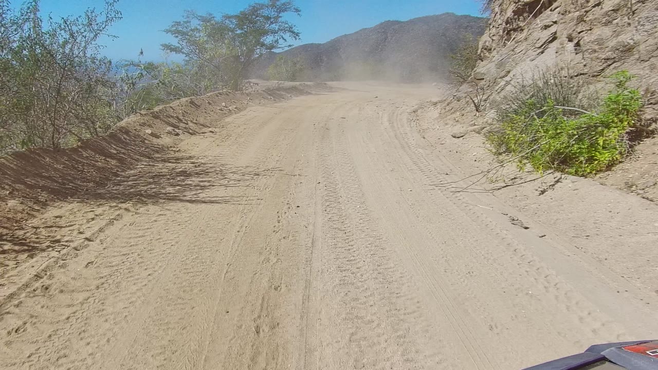 The Los Cabos trail curves through dry scrub and rock, with a dusty road surface, distant mountain ridge, clear sky, and no visible people.
