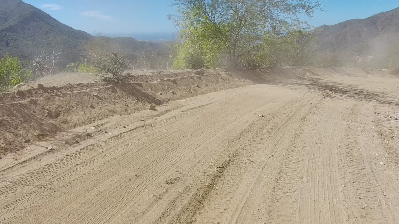 Start of a dusty dirt road in Los Cabos, Baja California Sur, with tire tracks, low bushes, mountains on both sides, blue sky, and no visible people.