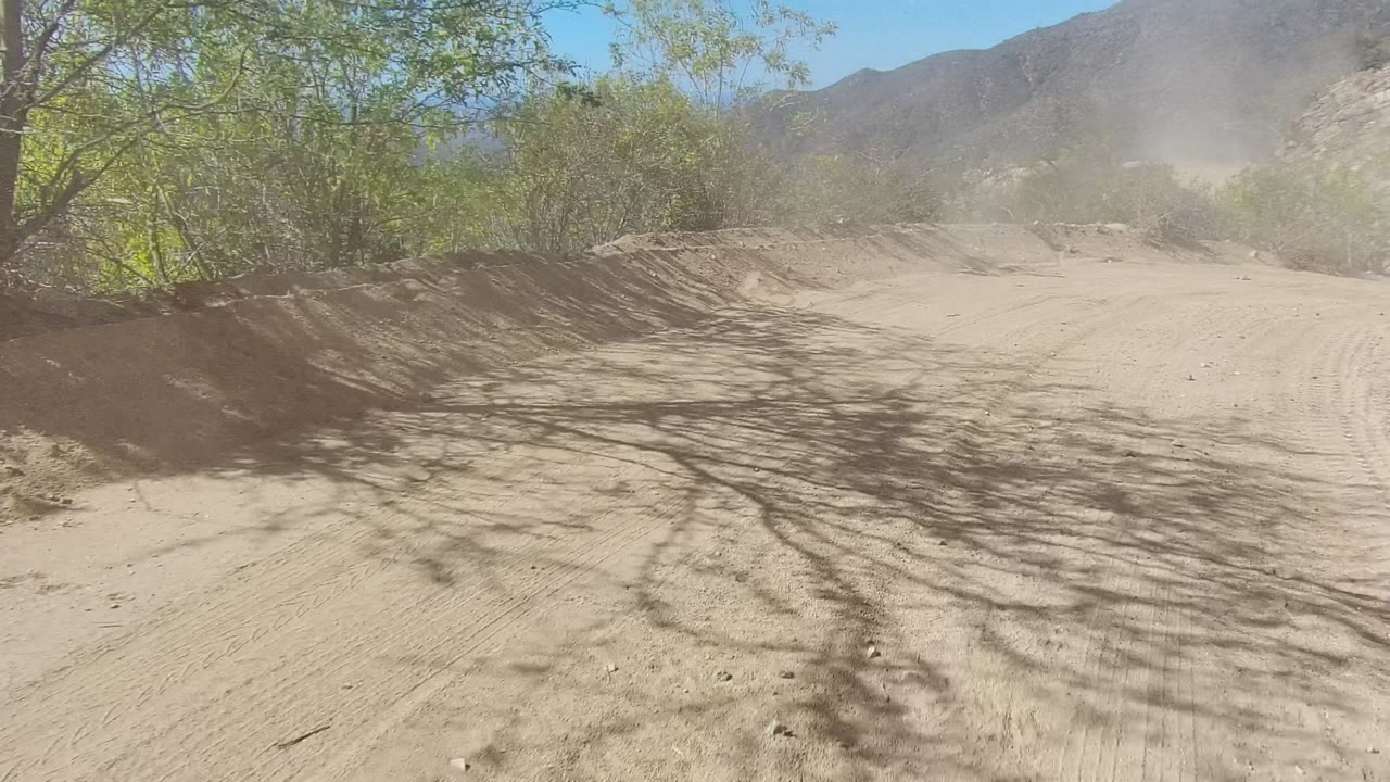 Sandy road surface with bushes, tree shadows, dust, and mountain ridges.