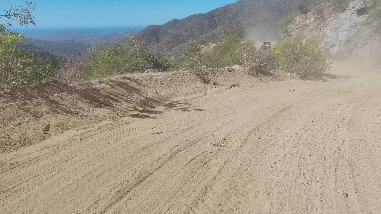 A bend in the Los Cabos dirt road passes scrubby green bushes, with loose dust hanging over the track, rocky hills behind, blue sky overhead, and no visible people.