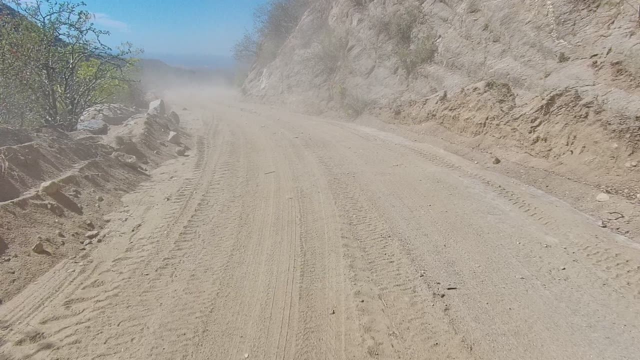 A dusty dirt road in Los Cabos, Baja California Sur runs beside a rocky hillside with dry brush, tire tracks, distant haze, and no visible people.