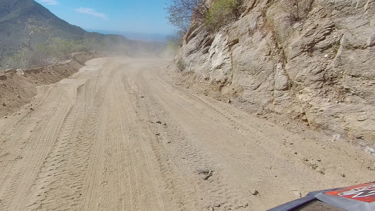 A red vehicle hood edge enters the lower frame as the Los Cabos dirt road follows a rocky wall with shrubs and dust ahead, with no visible people.