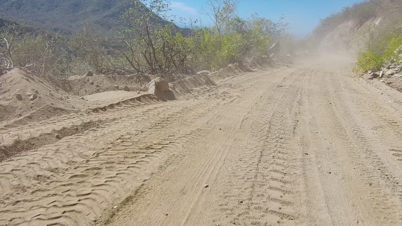 Sandy road curving through brush with a dust cloud ahead.