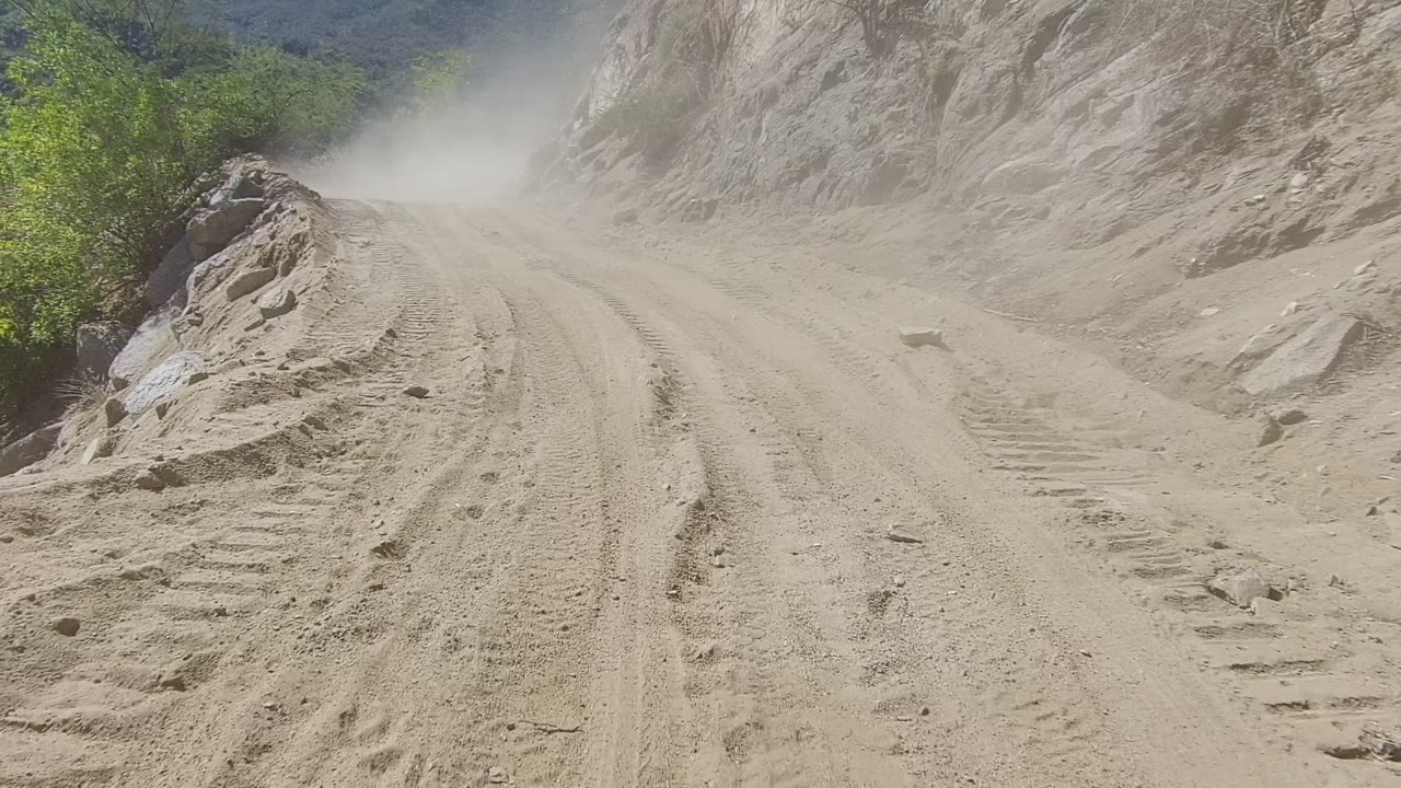 Dusty mountain dirt road in Los Cabos with deep tire tracks, loose rocks, sparse greenery, and no visible people.
