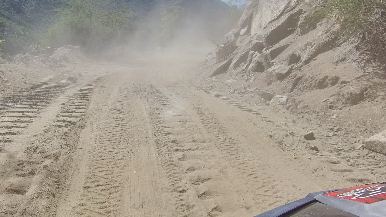 Rocky dirt road in Los Cabos with heavy tire impressions, dust drifting across the path, and a partial red vehicle edge at the lower right with no visible people.