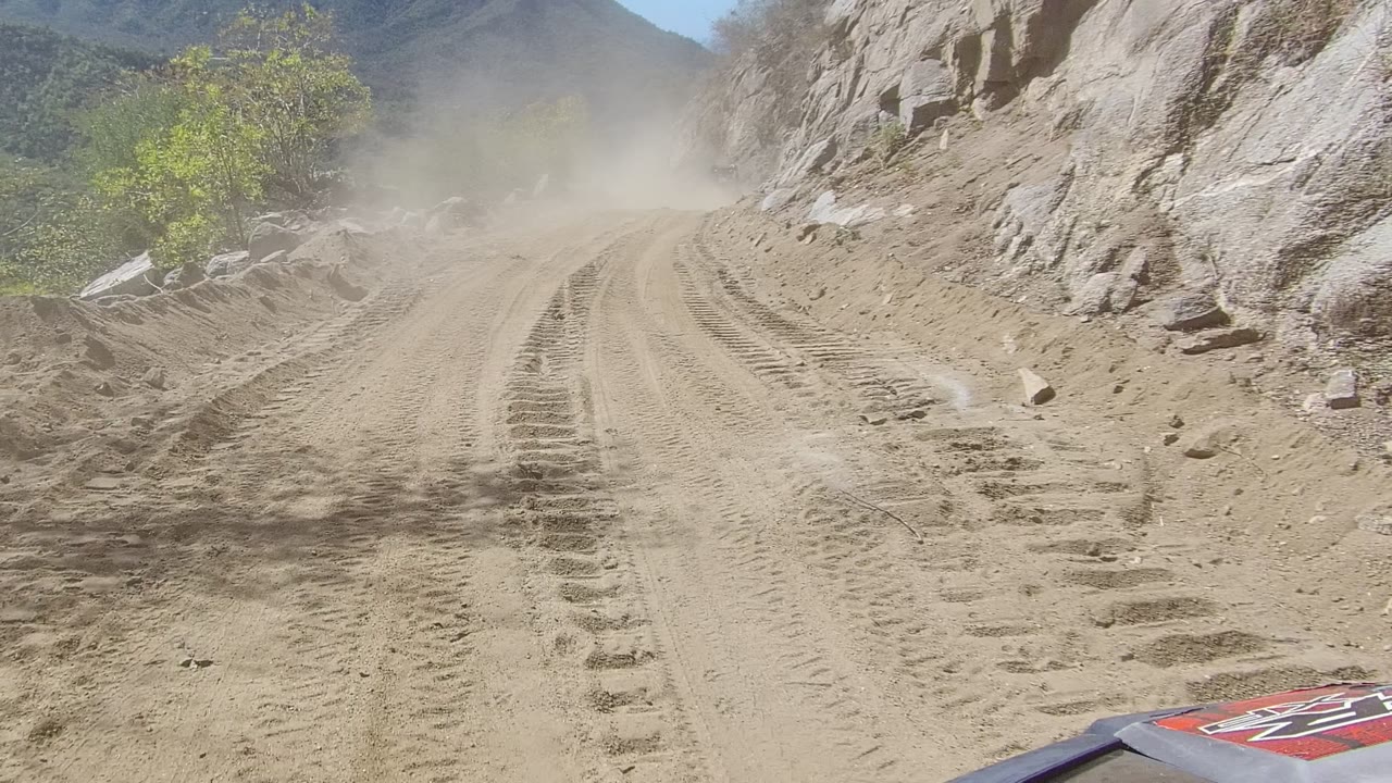 Dust hangs over a rutted dirt road in Los Cabos, Baja California Sur, with rocky slopes, sparse green trees, and no visible people.