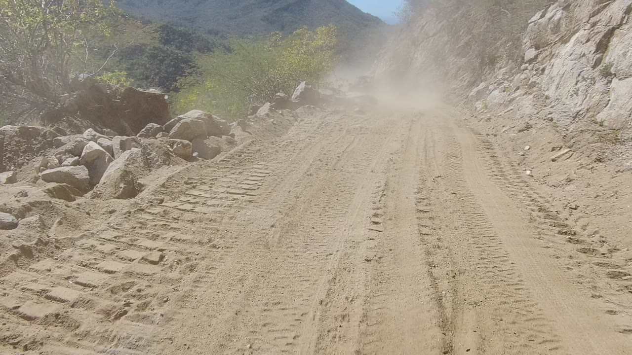 The dirt road in Los Cabos bends slightly through rocky terrain, with boulders along the right shoulder, green brush, lingering dust, and no visible people.