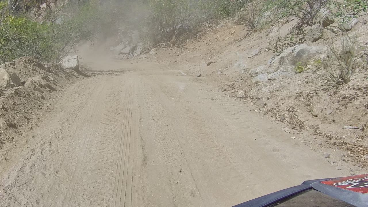 Opening frame of a dusty dirt road in Los Cabos, Baja California Sur, with tire tracks, rocky banks, sparse brush, a vehicle edge at the bottom, and no visible people.