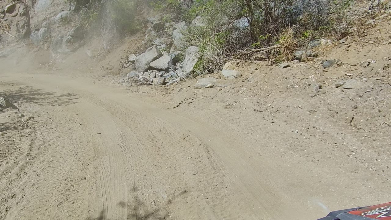 Dust hangs over the mountain dirt road in Los Cabos, Baja California Sur, with pale soil, rocks, brush, and the vehicle corner visible at the lower edge, with no visible people.