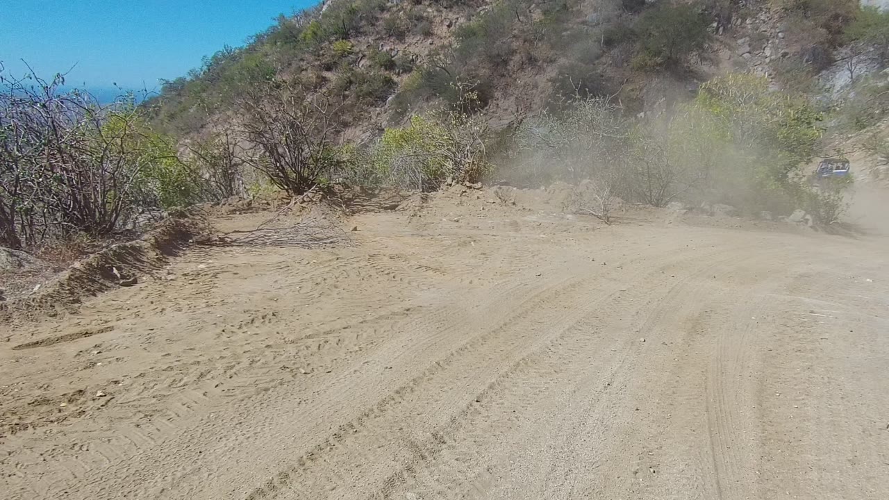 A sandy dirt road in Los Cabos, Baja California Sur curves through dry hills with tire tracks, brush, rocks, bright sun, and no visible people.