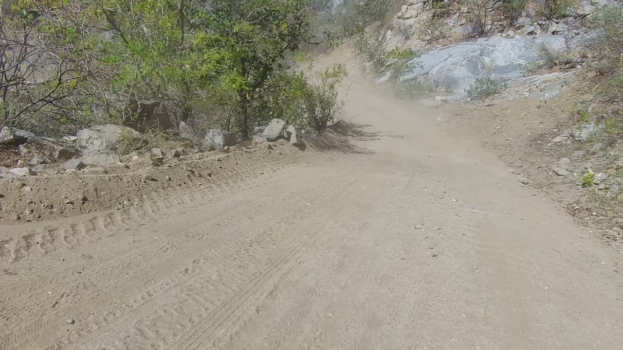 A dusty dirt road in Los Cabos, Baja California Sur passes pale rock outcrops and green scrub, with drifting dust and no visible people.