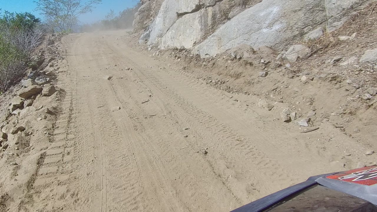 A sandy dirt road in Los Cabos runs uphill beside pale rock faces and scrub under a clear blue sky, with no visible people.