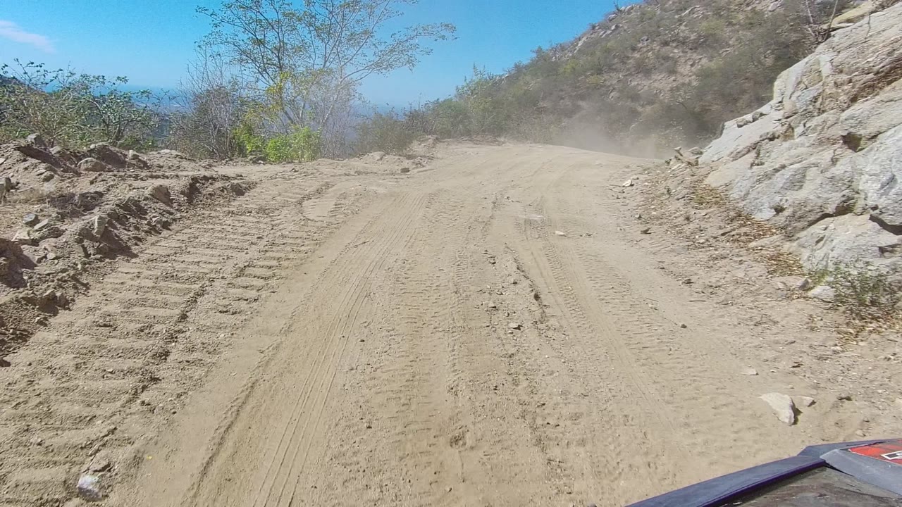 The road passes a scraped sandy shoulder and pale rock wall in Los Cabos, with dust hanging ahead and no visible people.