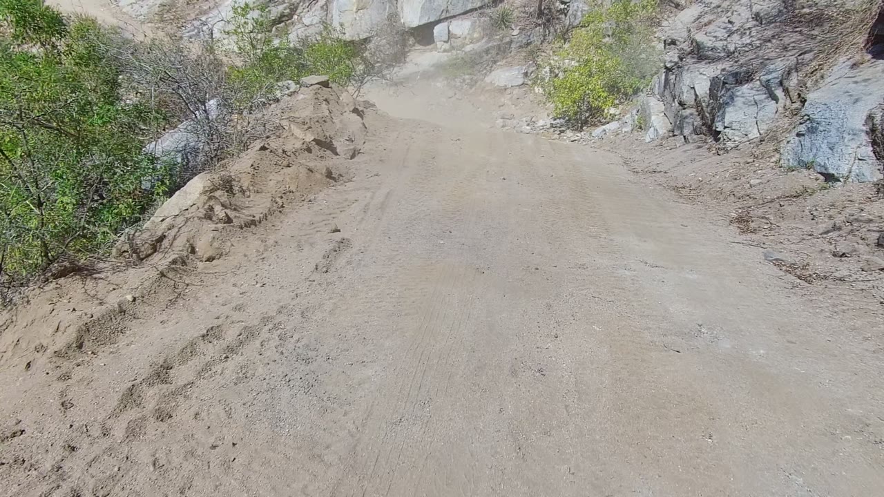 Opening view of a narrow dirt road in Los Cabos, Baja California Sur, with sandy berms, gray rock walls, sparse shrubs, and no visible people.