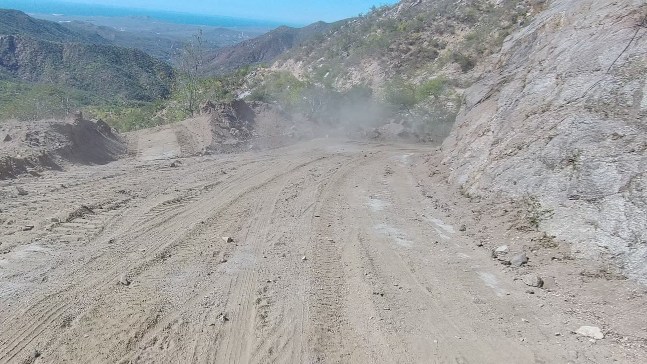 Dusty dirt road in Los Cabos, Baja California Sur, with tire tracks, rocky slopes, distant hills, sparse scrub, and no visible people at the start of the clip.