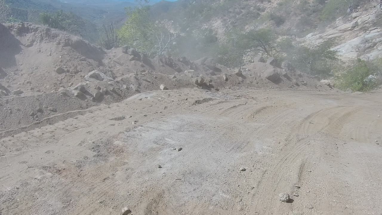 Rutted dirt track in Los Cabos with dust hanging over the road, rocky berms on both sides, scattered shrubs on the slopes, and no visible people.