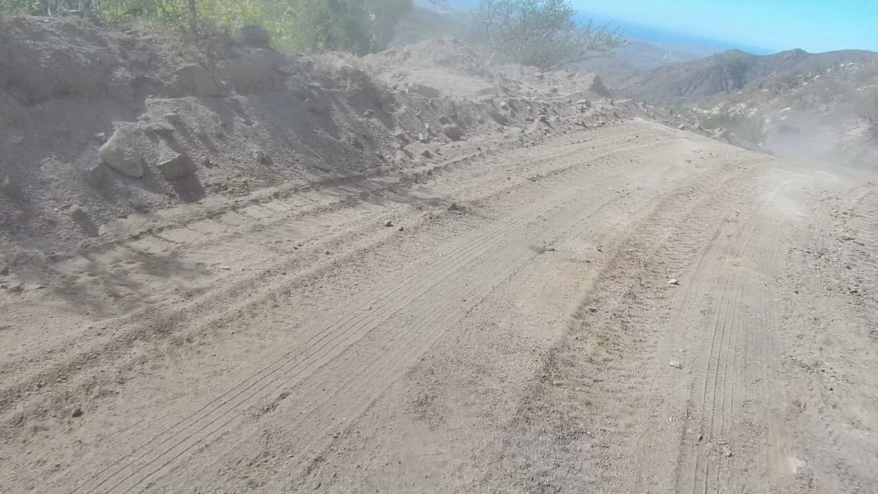 A dusty dirt road in Los Cabos, Baja California Sur runs between rocky slopes with tire tracks, sparse shrubs, and no visible people.