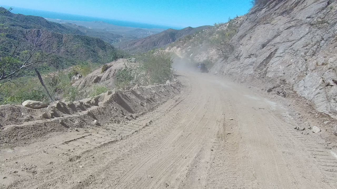 A curving dirt road in Los Cabos, Baja California Sur follows a rock wall with drifting dust, scattered stones, dry brush, and no visible people.
