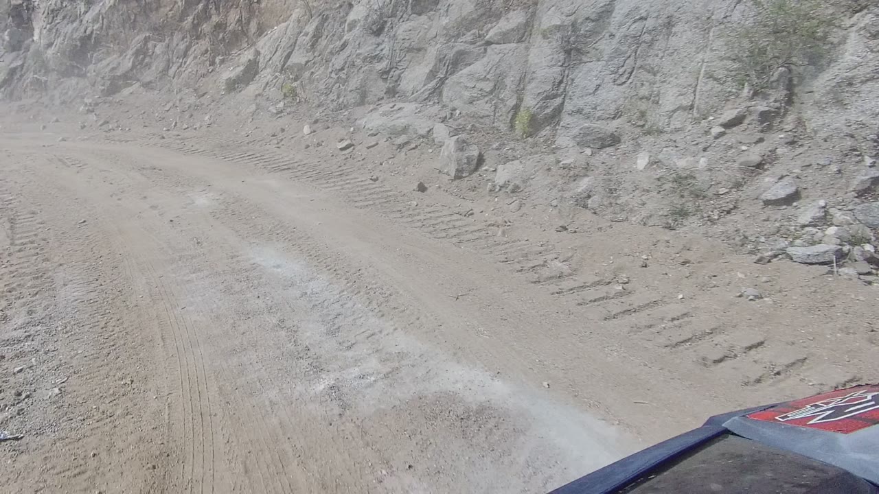 Opening view from a vehicle on a dusty Los Cabos dirt road bordered by gray rock cliffs, with part of the vehicle hood visible and no visible people.